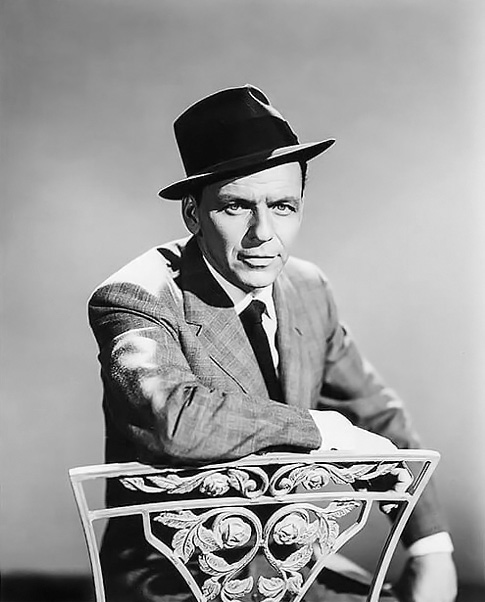 A man in a suit and hat sits backwards on a decorative chair, looking directly at the camera in black and white, evoking the timeless elegance of the Nat Sherman Townhouse.