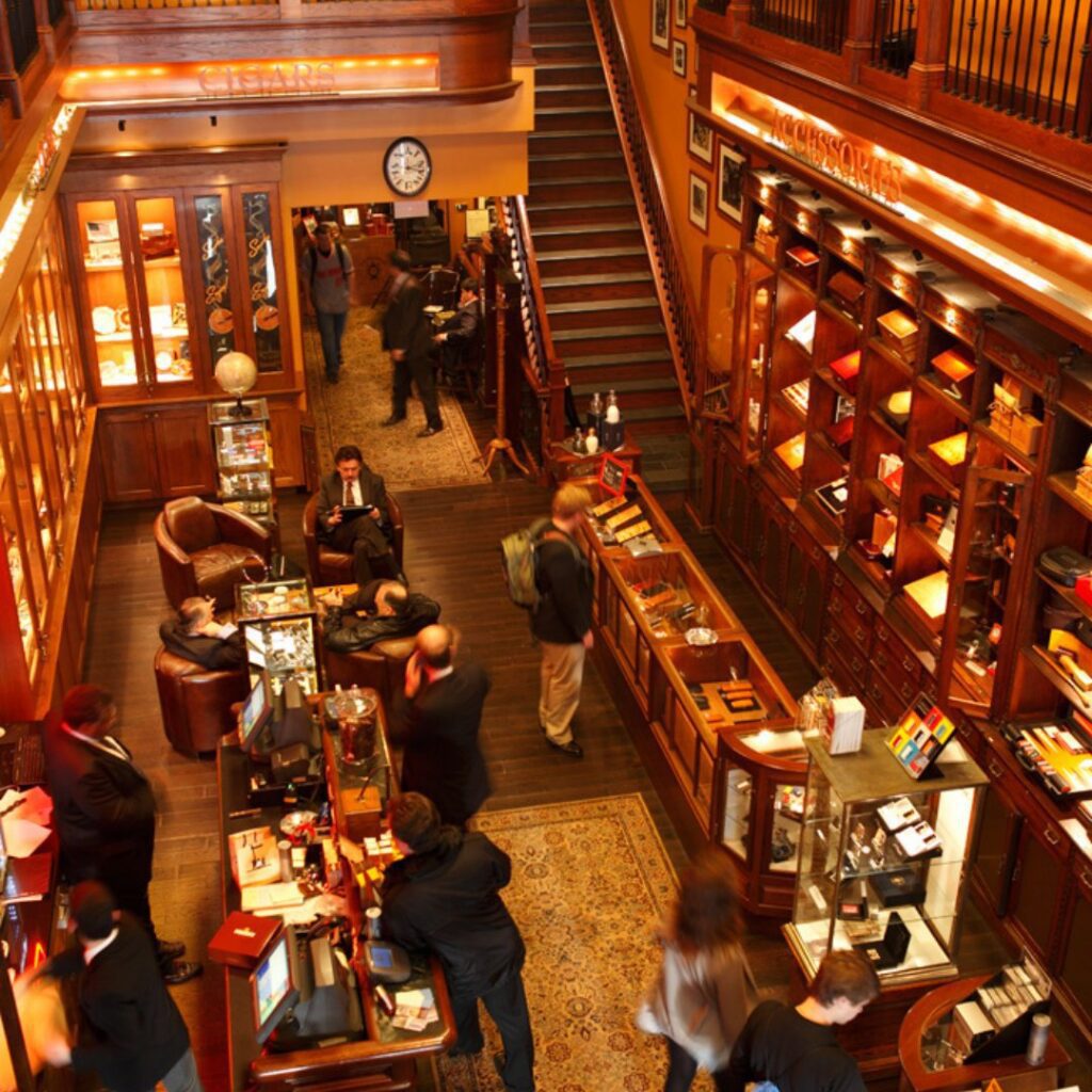 A busy, elegant shop with wooden displays, people browsing, and a staircase in the background captures the charm of the Nat Sherman Townhouse.