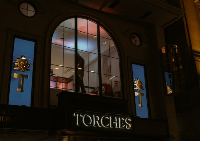 A person stands inside a lit window above a storefront with the sign “TORCHES” at night.