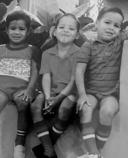 Three young children sit together on a ledge, smiling and wearing shorts and tall striped socks.