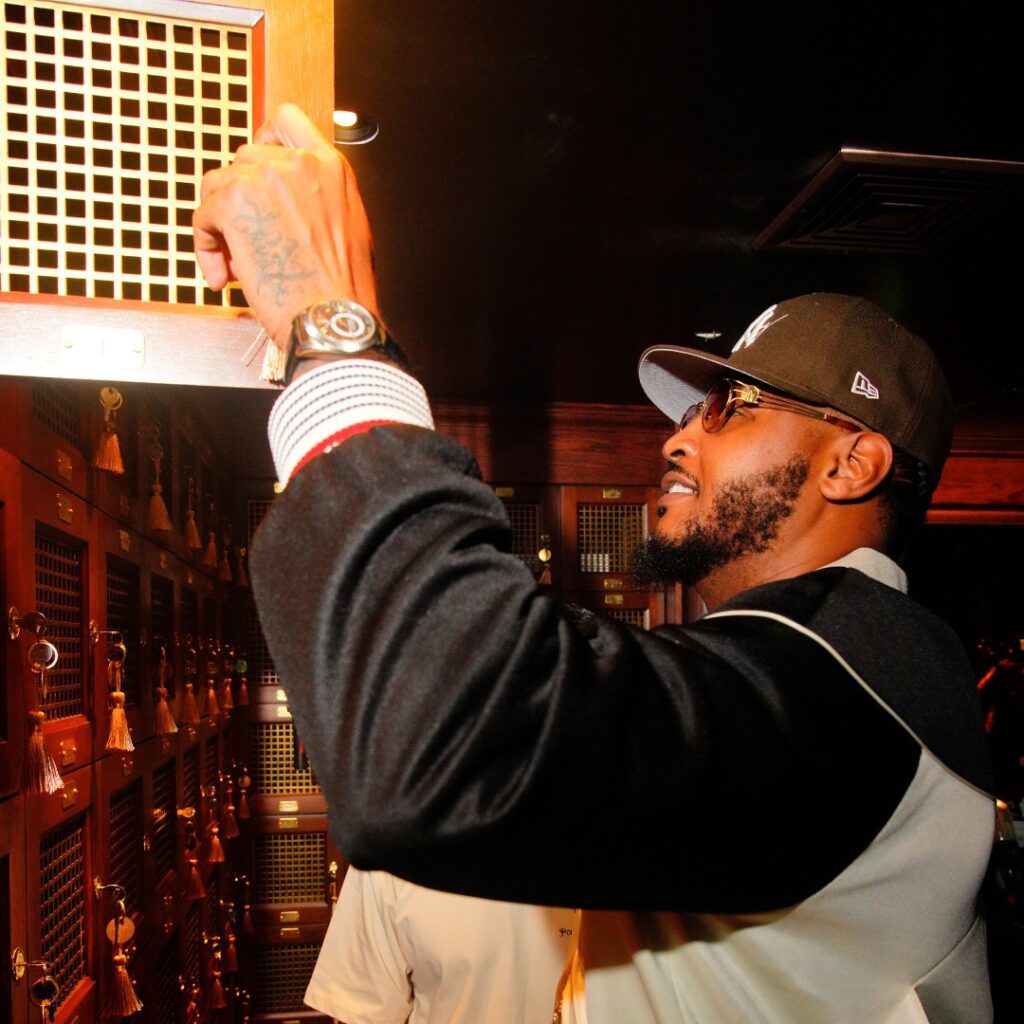 A man in a cap and sunglasses opens a locker in a dimly lit room with wooden lockers, gold key tags, and a subtle STAYME7O vibe.