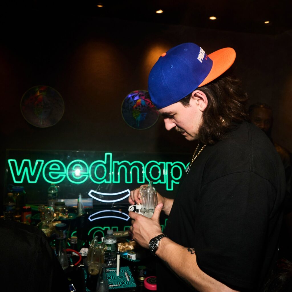 A man in a blue and orange cap holds a glass bottle at a table, with a neon weedmaps sign and a bold STAYME7O logo glowing behind him.