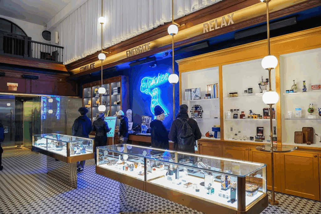 People browse glass display cases and shelves in a modern, brightly lit store with a neon sign, reminiscent of the vibrant atmosphere found at the High Times Cannabis Cup.