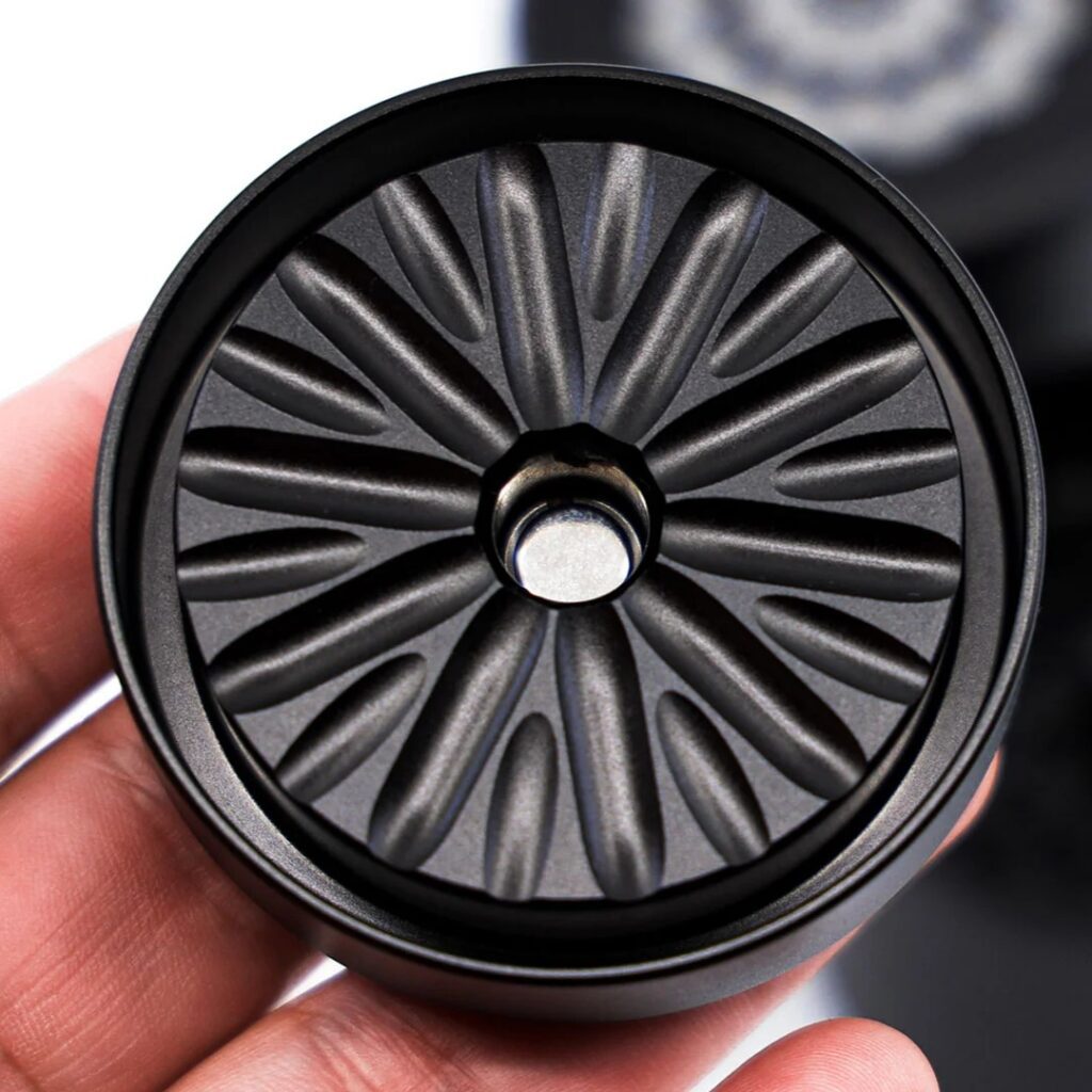 A hand holds a circular metal object with a radial, flower-like pattern inside, resembling a Flower Mill Grinder.