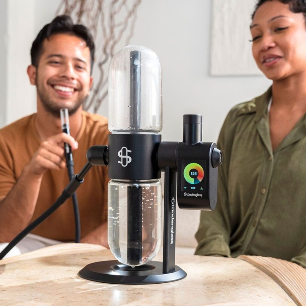 Two people smiling and sitting near a modern tabletop hookah with a digital display and water base.