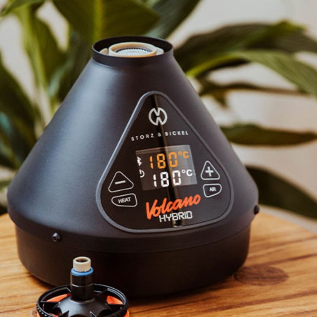 A Storz and Bickel Volcano Hybrid vaporizer displaying 180°C, placed on a wooden surface with plants in the background.