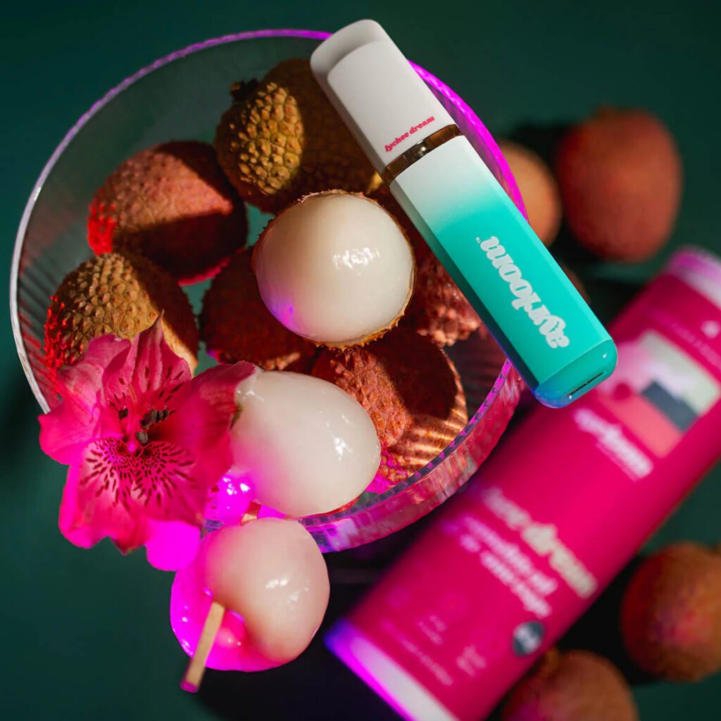 A glass bowl of lychees with a vapes device and a red drink can, beside a pink flower, on a green surface.