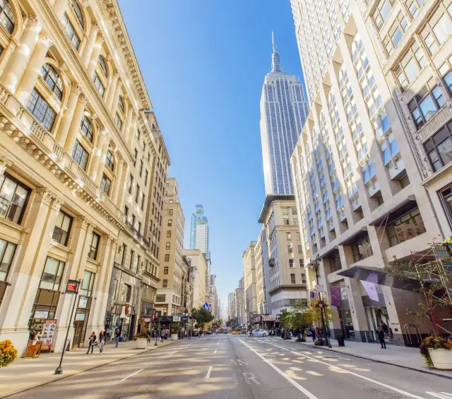 Wide street in New York City with tall buildings and the Empire State Building in the background under a blue sky.