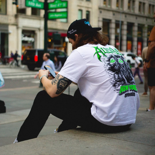 Person with long hair and tattoos sits outdoors on a ledge, wearing an Alien Labs Brand graphic t-shirt and looking at their phone.