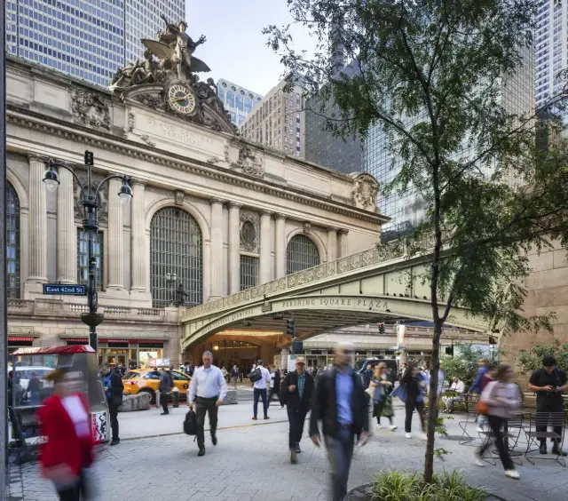 People walking outside Grand Central Terminal in New York City, with skyscrapers and trees in the background.