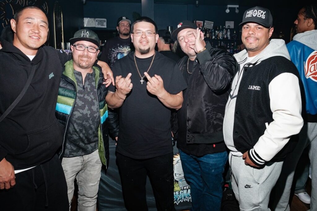 Five men pose together and smile inside a busy, dimly lit bar or club, their laughter as vibrant as the juicy fruit strain.