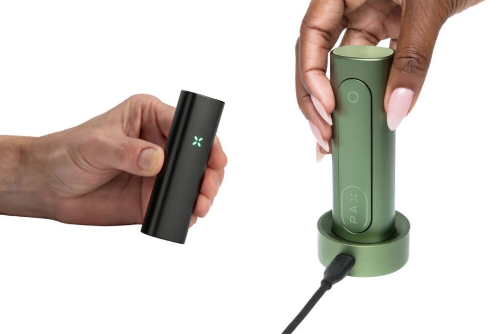 Two hands hold electronic vaping devices, one black and one green, with the green device in a charging dock.