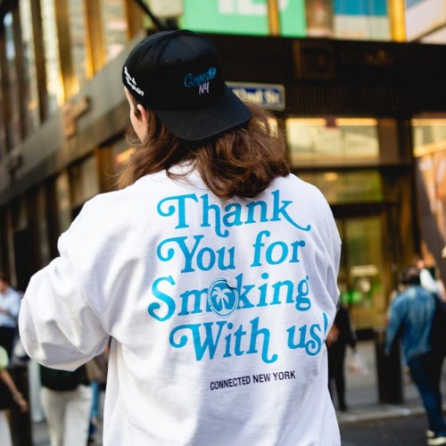 Person wearing a white sweatshirt with Thank You for Smoking With us! and Connected Cannabis branding on the back, standing on a city street.