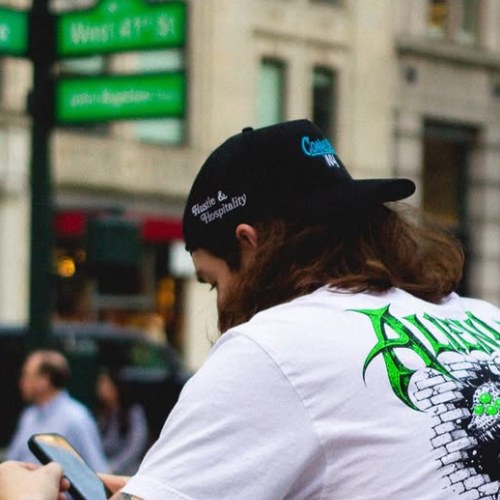 Person in a white graphic tee and black cap looks at phone on a city street, green street signs in the background—staying connected while repping their favorite Connected Cannabis brand.