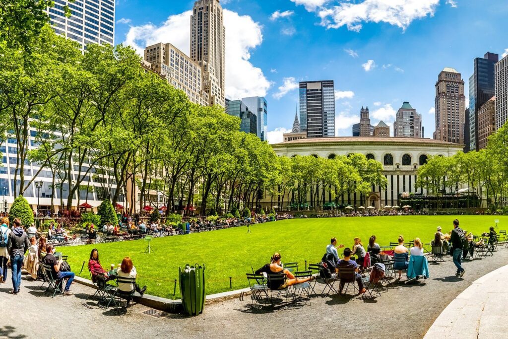 People relaxing on chairs around a green lawn in a city park surrounded by tall buildings on a sunny day, just steps from the Bryant Park Dispensary.