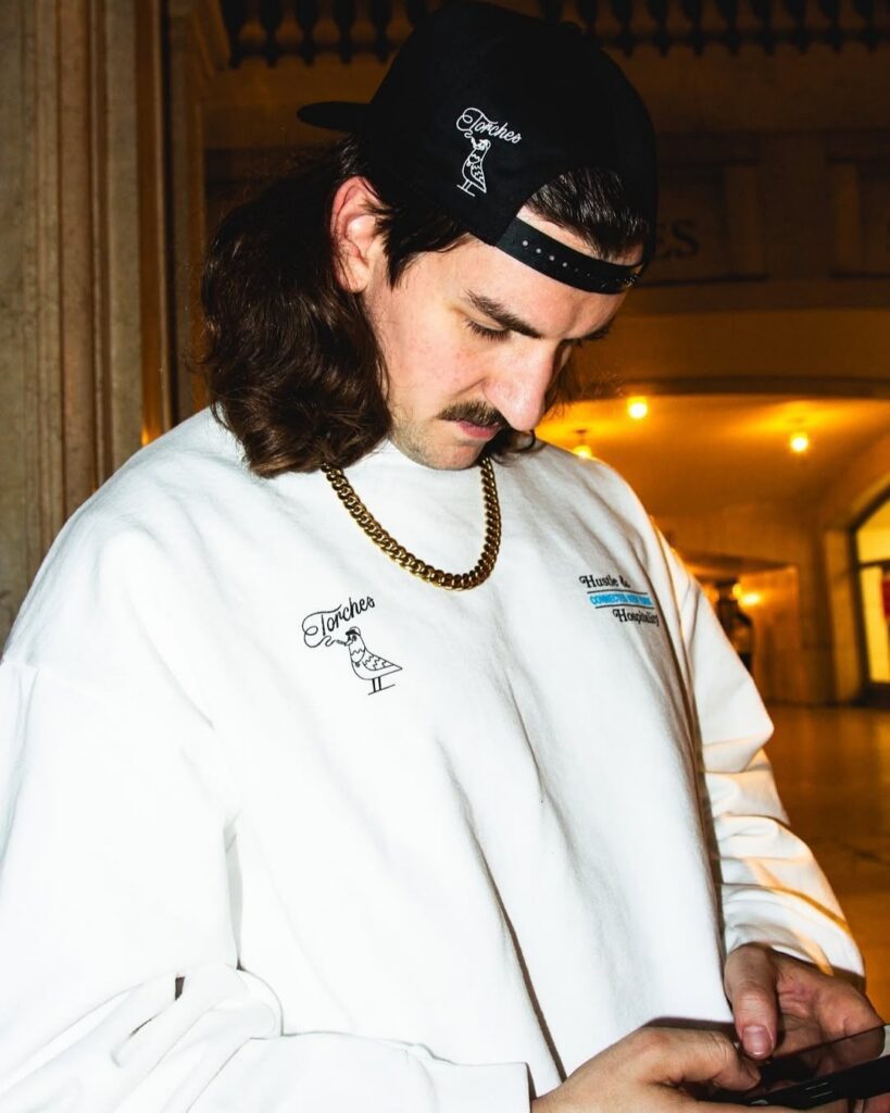 Man in a white sweatshirt, gold chain, and black cap looking down at his phone, perhaps checking out the latest from Connected Cannabis indoors at night.