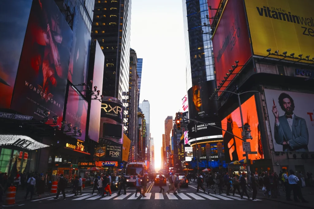 Crowded Times Square at sunset with bright billboards, busy crosswalk, and people walking in all directions.