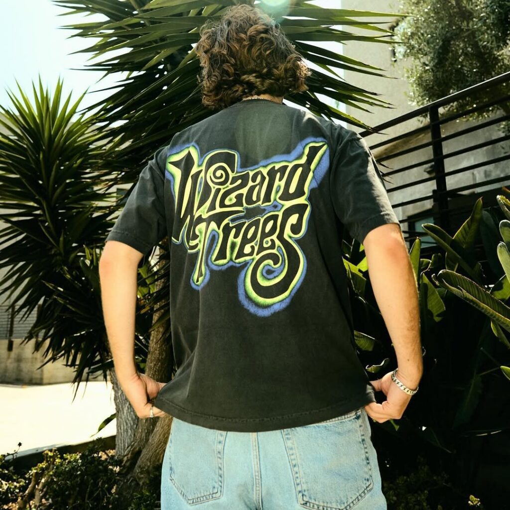 Person wearing a “Wizard Trees” t-shirt stands outside among green plants with their back to the camera.