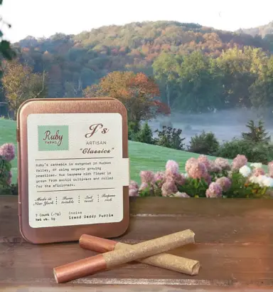 A tin of pre-rolls from Torches NYC sits on a table outdoors with three joints and a scenic, misty landscape in the background.