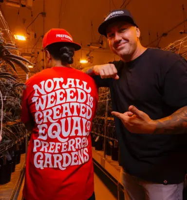 Two people in a grow room, one has a red shirt saying, Not all weed is created equal. Preferred Gardens. Bright lights reminiscent of Torches NYC illuminate the lush plants around them.