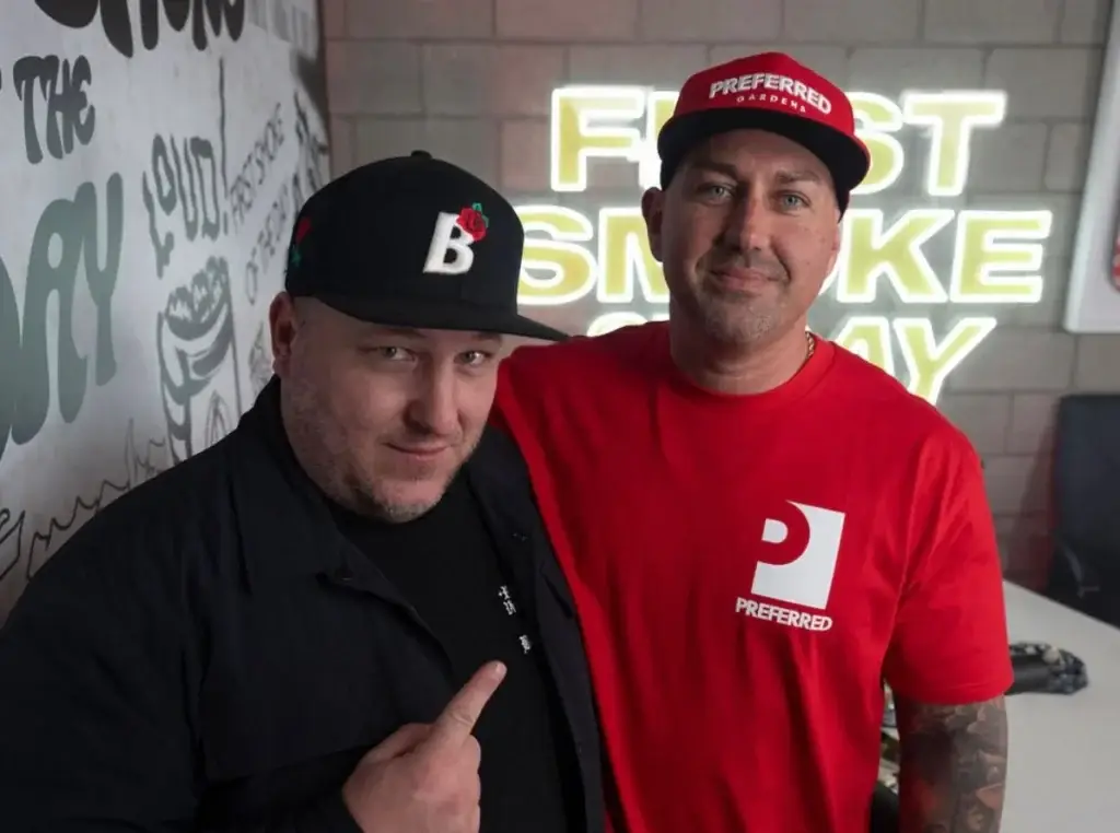 Two men posing together indoors; one wears black, the other wears red and a Preferred cap, with neon sign behind.