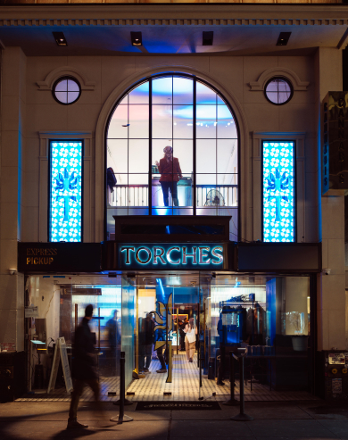 Nighttime view of a restaurant entrance with neon TORCHES sign and people visible inside and outside.