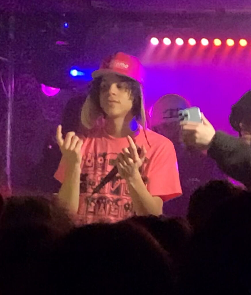 Person in a pink shirt and hat performs on stage with colorful lights in the background at the Weedmaps Kickback.
