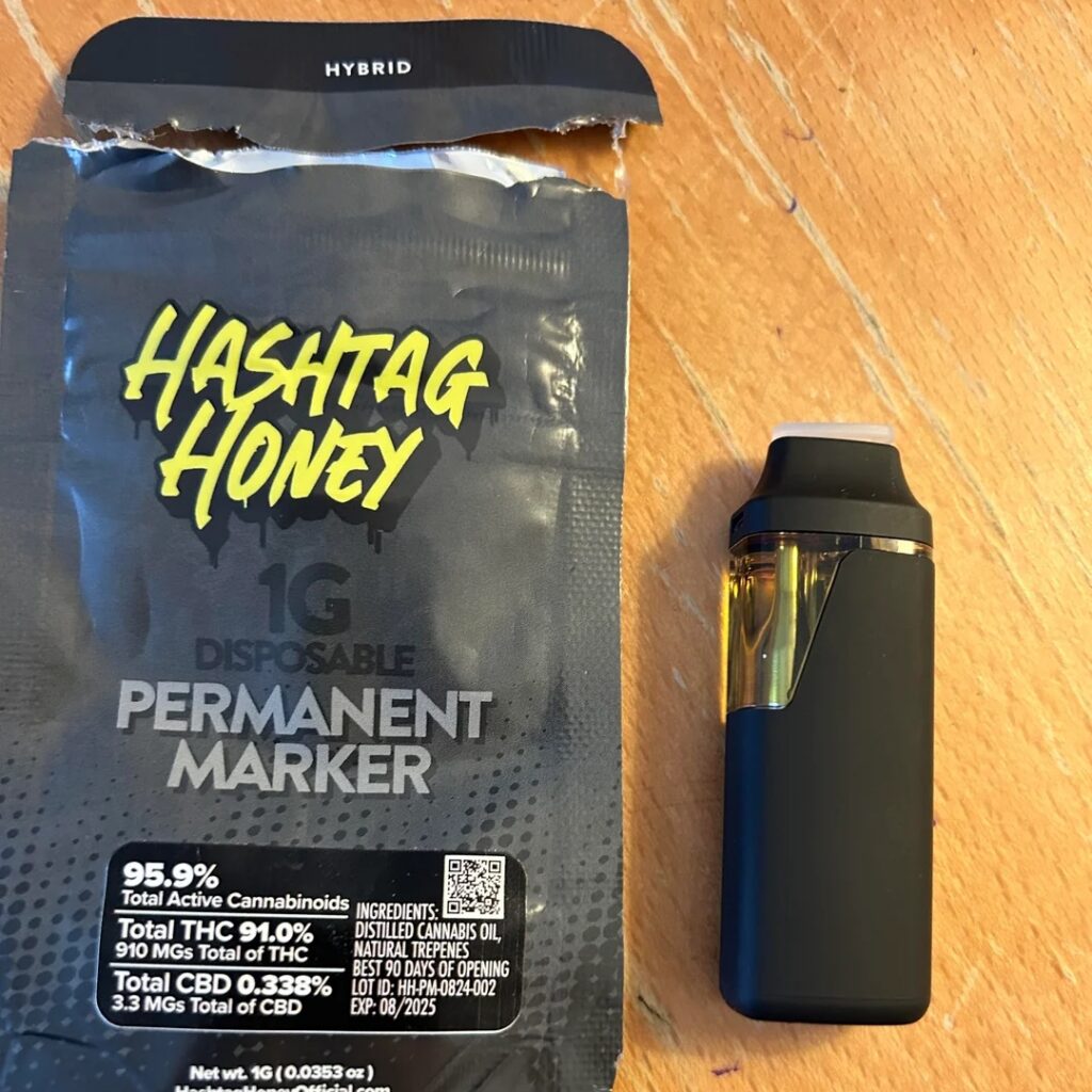 Black vape pen beside Hashtag Honey hybrid disposable packaging with yellow text on a wooden surface.