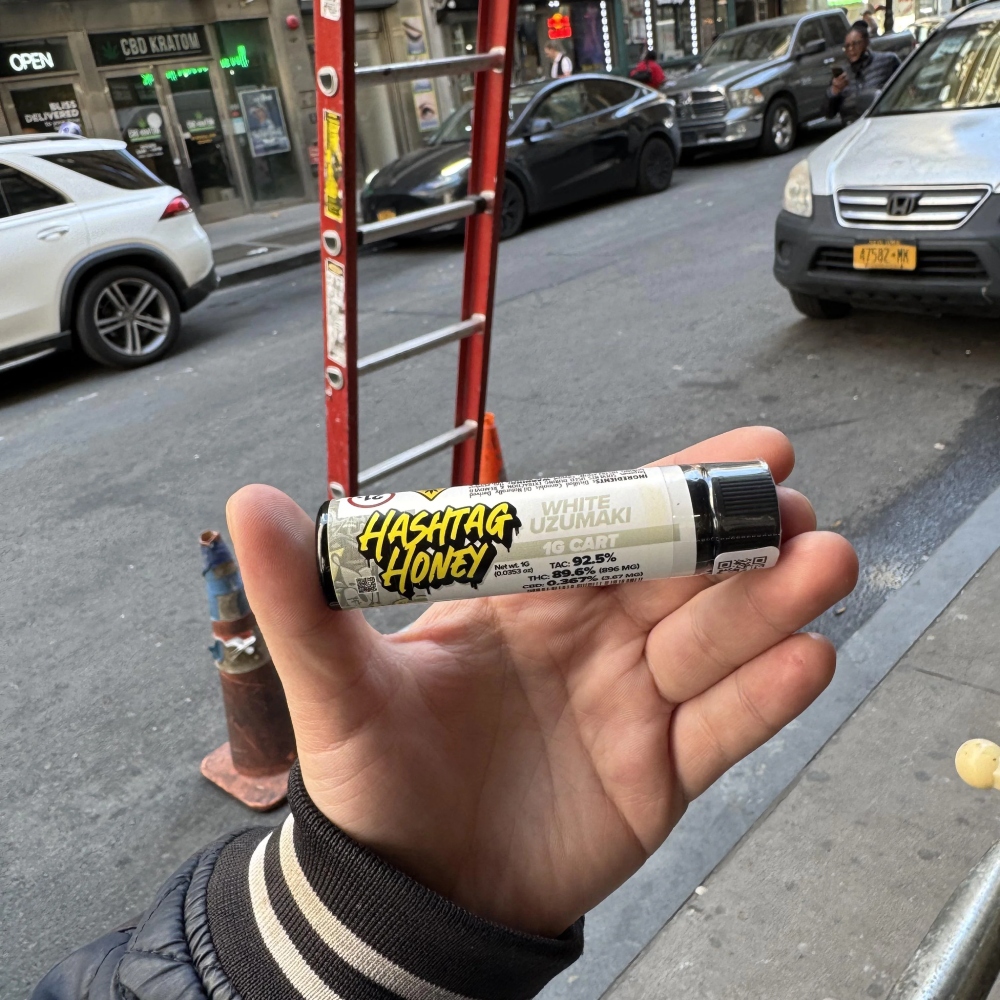 A hand holds a Hashtag Honey cannabis pre-roll on a city street with cars and shops in the background.